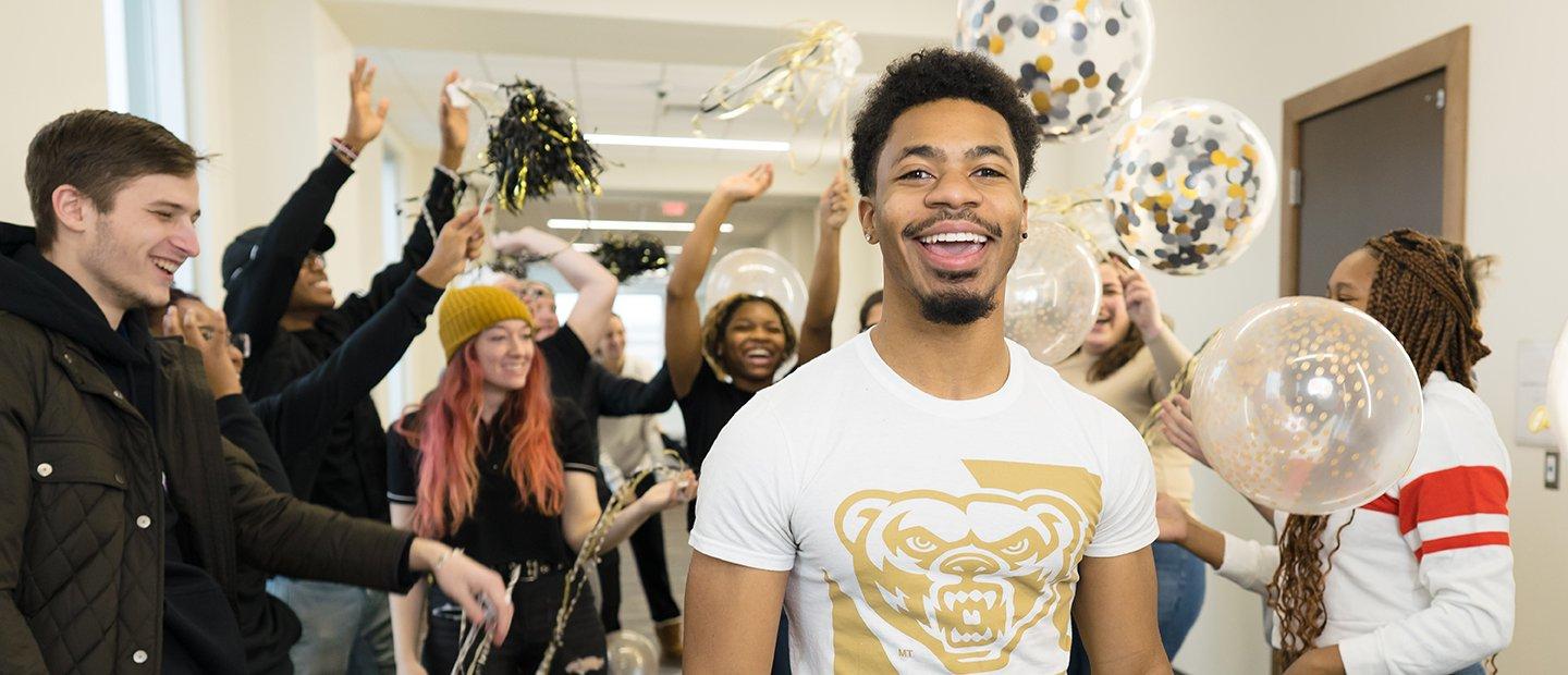 A young man smiling with a crowd behind him, holding black and gold balloons and ribbons.